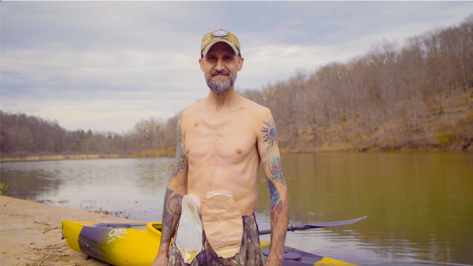 a man with a beard and tattoos standing by a body of water